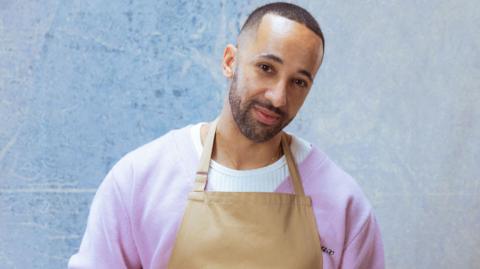 A man with short black hair and beard stands with his head cocked to one side. He is wearing a purple cardigan over a white jumper, with a brown apron over the top.