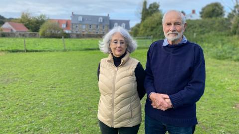 An older couple with grey hair stand in a field, with a several houses, a hedge and a post and wire fence behind. She wears a cream body warmer and black top and trousers, plus glasses. He is wearing a navy blue guernsey-style woollen sweater, jeans, and a blue shirt and has white hair and is bald on top. He clasps his hands.