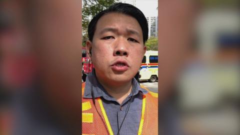 A man wearing an orange jacket over a shirt, with an ambulance and fire engine behind.