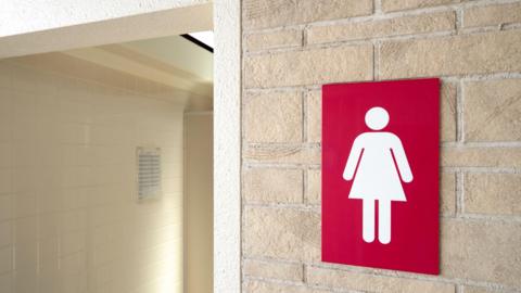 A generic photo of a ladies' toilet sign on a wall outside a toilet. The sign has a white lady symbol on a red background. The doorway has pale cream tiles.