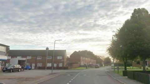 Sherlock Avenue in Haydock with shops on the left and trees on the right. Two blocks of two-storey terraced houses are in the distance at right angles to each other.