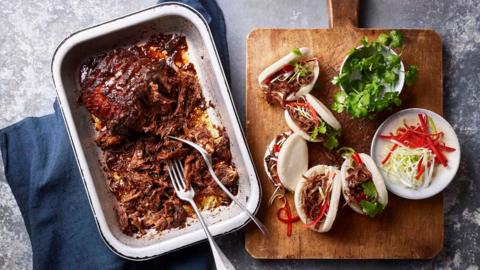 A roasting tray with pulled brisket alongside a chopping board of bao buns filled with the beef and sliced chillies and lettuce to go inside