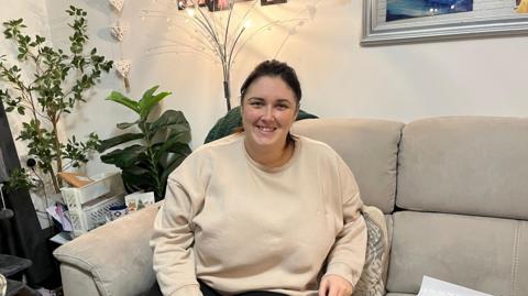 A woman in a cream jumper sitting on a grey sofa smiling. Behind her is a decorative lamp along with two house plants. There's also a coffee table to the left of her with a mug and various stationary bits tucked in.