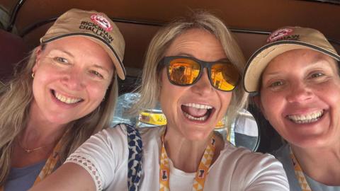 Three middle aged women with blonde hair in the back of a tuk-tuk. Two are wearing caps and another is wearing black-framed sunglasses with reflective orange lenses. All of them are smiling.