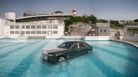 A black Rolls Royce Phantom car in an outdoor swimming pool shot from directly above on a sunny day. The lido building behind sits below Plymouth Hoe, with its iconic red and white lighthouse behind. 