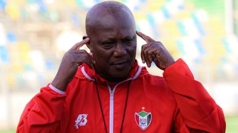 Kwasi Appiah, wearing a red tracksuit top with a Sudan FA logo on the left breast, points his index fingers to his temples