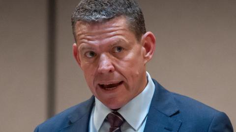 Rhun ap Iorwerth speaking wearing a blue suit, blue shirt and a red tie
