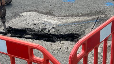 A hole in the middle of the road. Orange closure barriers are visible in the foreground. A machine can be seen poking at the hole on the left.