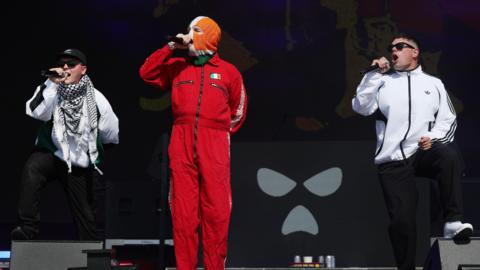 The band Kneecap perform onstage - all three members of the band are holding microphones. One of the group, standing centre, is wearing all red with a small Irish flag printed on it and a tricolour balaclava. The other two band members are wearing white jackets and sunglasses.