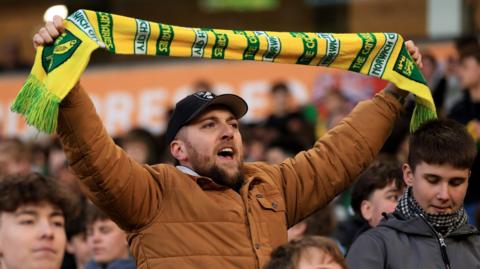 Supporter wearing a brown jacket and black baseball cap holds a green and yellow Norwich City scarf above his head during their FA Cup tie against West Bromwich Albion