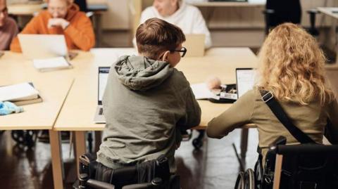 Several students are seated around rectangular tables in a classroom, each using a laptop. Two students in the foreground are seated in wheelchairs, positioned side by side at the table, viewed from behind. Other students are seated further along the table, also working on laptops. The room has a polished floor, light‑coloured tables and chairs, and computer monitors visible in the background.