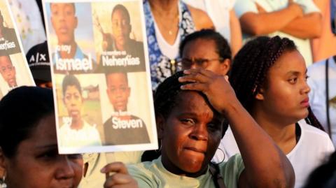 A person reacts during a tribute dedicated to Ismael and Josue Arroyo, Nehemias Arboleda and Steven Medina in Guayaquil on 8 December 2025. The woman clutches her forehead while holding up a composite picture of the four boys. Around her are other women also taking part in the tribute. 