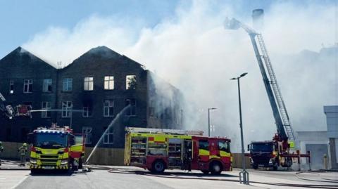 Three fire engines are outside a burning building, which looks like an old mill and is missing a roof 