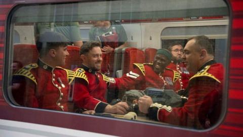 A group of guards sitting on a train with red seats. They are wearing the red uniform with yellow shoulders. They are smiling and chatting, one man has his had on the shoulder of another.