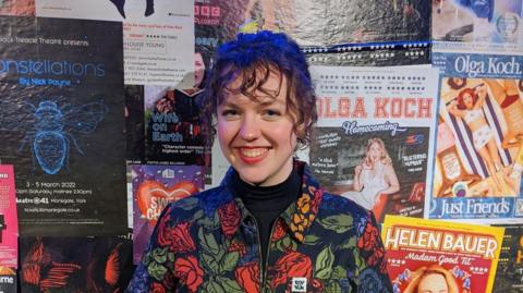 Elspeth Snelling, a young woman, stands against a backdrop of theatre and comedy posters. She has dark wavy hair pulled back from her face. She wears a collared dark floral jacket and black polo neck jumper. She is smiling. 