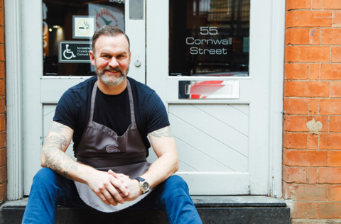 A man with dark hair and a beard in a dark tshirt and jeans, sat on a step in front of a white door