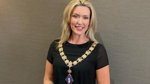 A woman, Kerry Burley, smiles at the camera. She has shoulder length blonde hair and is wearing a large gold chain which has a pendant with Robert Burns's face on it.