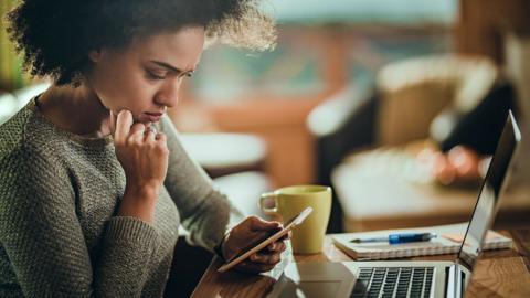 A woman looks at her phone while frowning with a concerned look on her face, as she sits at a desk at home with a laptop, coffee cup and notepad and pen in view