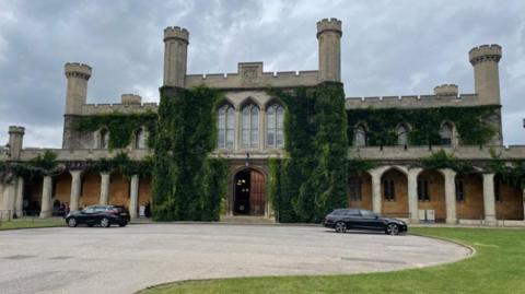 An exterior shot of Lincoln Crown Court which is a Gothic style building with turrets, greenery coming from it and large windows and a wooden front door. Two black cars are parked in front.