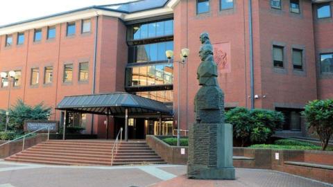 The courtyard outside the front entrance to Birmingham Crown Court. A statue with multiple heads is on the right. Stairs lead up to the canopy above the main doors on the far side.