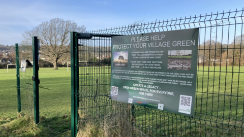 The entrance to Stoke Lodge field. there is a green gate with a large green poster reading 'please help protect your village green'.