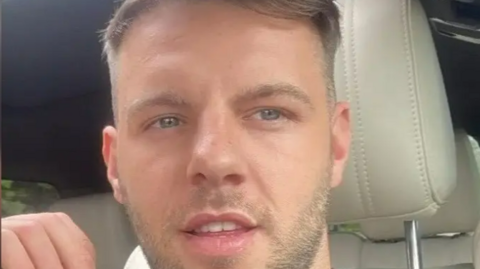 A selfie image taken by Jon Belfield shows him sitting in the front seat of a vehicle. He has short brown hair and brown stubble. 
