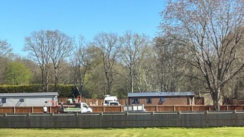 A field with fences in it. In the background, two mobile homes are surrounded by vans and diggers, with people working nearby. There are trees behind this, and a blue sky.