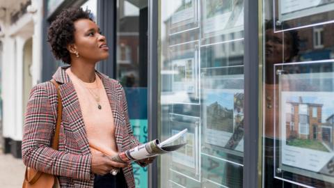 A stock image of a lady perusing an estate agent's available properties in the window. She carries a newspaper, a handbag, and a blazer.