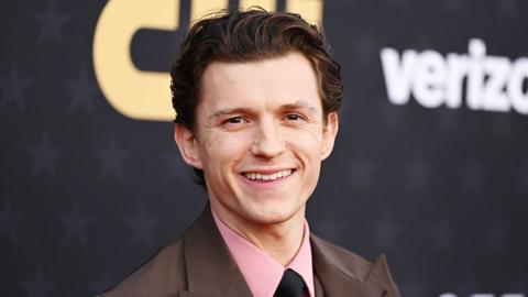 Male actor Tom Holland, wearing a pink shirt, black tie and brown jacket, smiling.