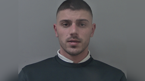 Man with dark short cropped hair and stubble. He is wearing a white shirt underneath a dark jumper.