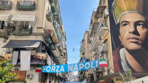 Naples alleyway between two buildings with a banner reading "Forza Napoli" strung between them and a large mural of Maradona on the side of one of the buildings