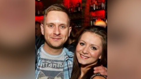 A blond man in a grey band t-shirt and a blue plaid shirt puts his arm around a pretty woman with long brown hair. 