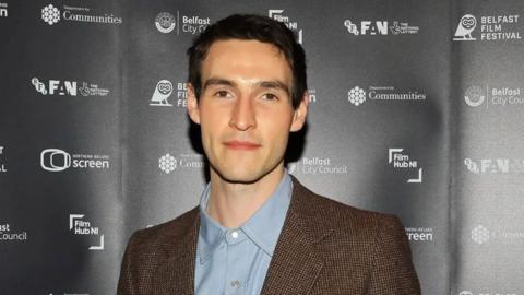 A man with short dark hair and eyebrows. He is wearing a blue shirt and brown suit jacket.