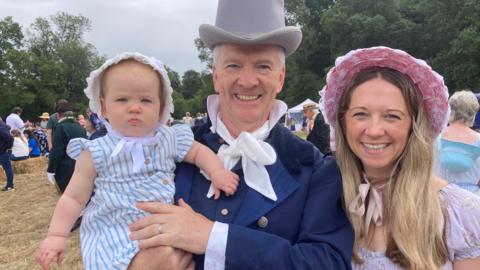 Baby, man and woman in Regency dress wearing bonnets and top hat at a Jane Austen celebration.