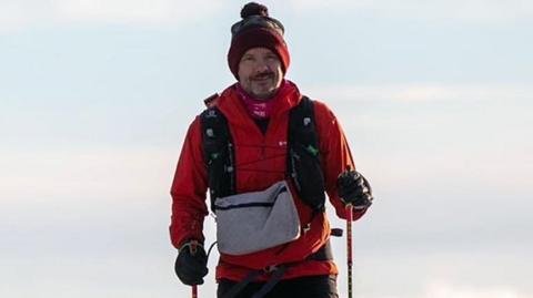 Stephen Chamberlain is shown in winter clothing, including a hat, and is seen wearing gloves and holding sticks to aid walking in the conditions