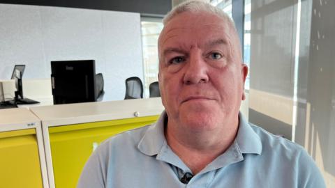 Simon Byrne with grey hair and wearing a grey polo top sitting in an office with a mustard coloured filing cabinet looks impassively.
