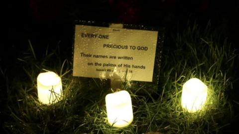 Three led candles on grass lit during a candlelight vigil at a cemetery to honour those buried in a mass grave with a yellow sign which reads: Every one precious to God: Their names are written on the palms of His hands. Isaiah 49 v 15 and 16 from the Bible 