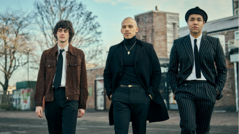 Three men, who are characters from This Town, walk in a line. The man on the left wears a brown suede jacket, a white shirt, black tie and black trousers. The man in the middle wears a black suit with a black turtleneck and a golden chain around his neck. The man on the right wears a black pinstripe suit, a black tie, and a black hat. They are walking on a street in front of a small redbrick building.