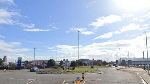 A roundabout in the middle of a dock junction, one car goes around to the left of it, it is a sunny day with blue skies