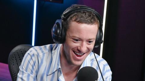 Joseph Quinn smiles in a Radio 1 studio - he wears headphones and a white and blue striped shirt