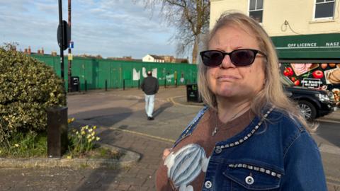 Linda Kincaid with long blonde hair wearing sunglasses and a blue denim jacket. She is standing in a public square with a tree to the left and an off licence with a green hoarding to the right. In the distance, there is a green hoarding around a demolition site.