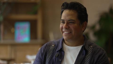 A man in a white t-shirt and navy blue over-shirt. He sits inside the restaurant, which is blurred behind him. 