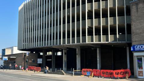 Exterior shot of the George Street car park in Hull. It is a 1960s Brutalist-style structure. Access to the car park has been blocked off with red traffic barriers. There is a person seated on the wall at the front of the facility.