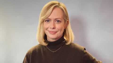 A woman with shoulder length blonde hair wearing a brown turtleneck.