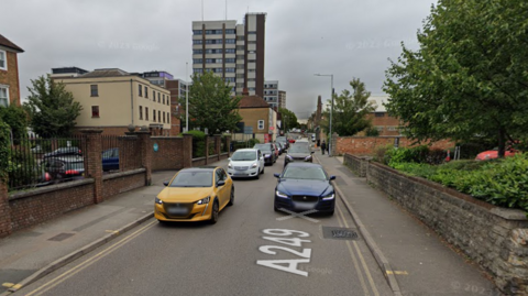 A one-way street with cars - one side is a stone wall and the other has black railings. In the distance there is a high rise building. 