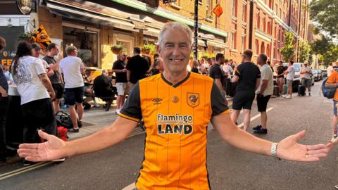 John Drinkall wearing an orange and black Hull City shirt. He has grey hair and is posing with his arms wide, while smiling at the camera. Fans are gathering outside a pub behind him.