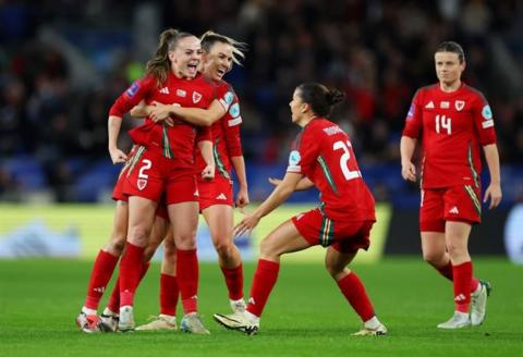 Wales women celebrating after their win to qualify for the Euros 2025. They are on a pitch and wearing a red kit. There are five of them and one is hugging another. 