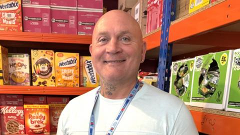 A man with a tattooed neck. He is wearing a white T-shirt and standing among rows of boxes of food, including cereals and crisps.