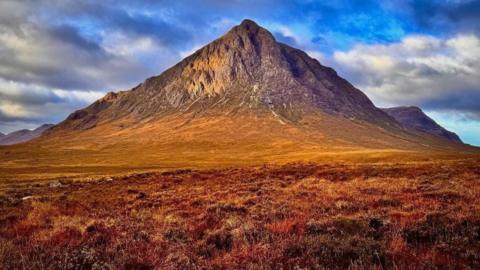 This sole mountain dominates the picture. It is rocky. In the foreground is grass and heather which is red and yellow in colour.