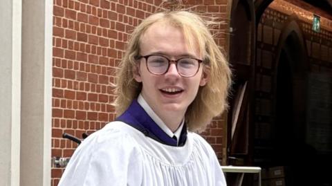 Alastair wears choir robes and has long blonde hair and glasses.